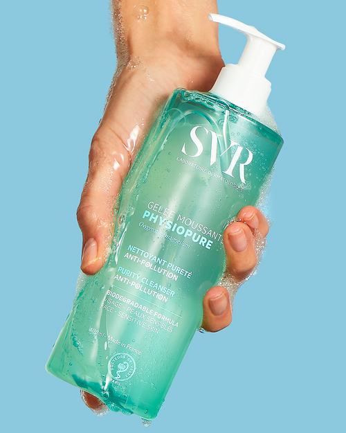 Hand hält eine türkisfarbene SVR PHYSIOPURE Gelée Moussante Flasche mit weißem Pumpspender und sanftem Reinigungsschaum.