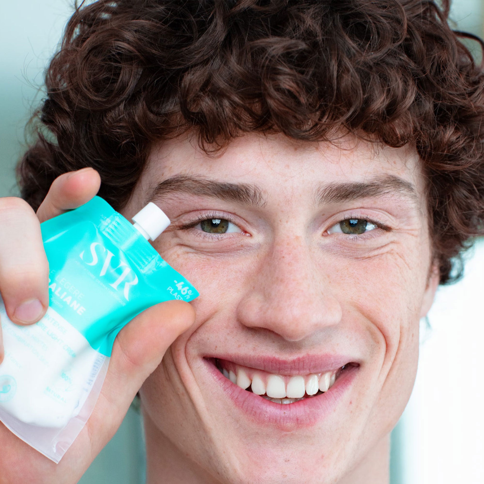 Junger Mann hält eine türkisfarbene SVR Feuchtigkeitscreme im praktischen Beutelformat an sein Gesicht.