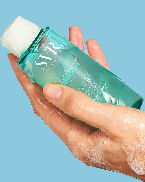 Hand hält eine türkisfarbene SVR PHYSIOPURE Gelée Moussante Flasche mit weißem Verschluss und schäumendem Reinigungsgel.