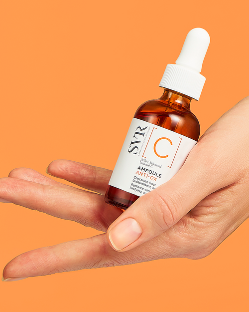 Hand hält SVR Ampoule Anti-Ox Serum in einer braunen Glasflasche mit weißer Pipette vor orangefarbenem Hintergrund.