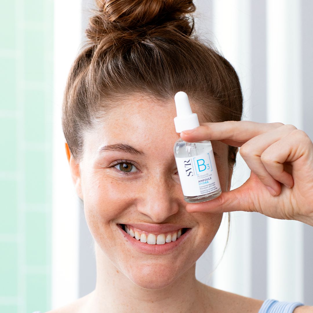Lächelnde Frau hält ein SVR Ampoule Hydra Serum mit Vitamin B3 vor ihr Auge für eine Feuchtigkeits-Boost-Routine.