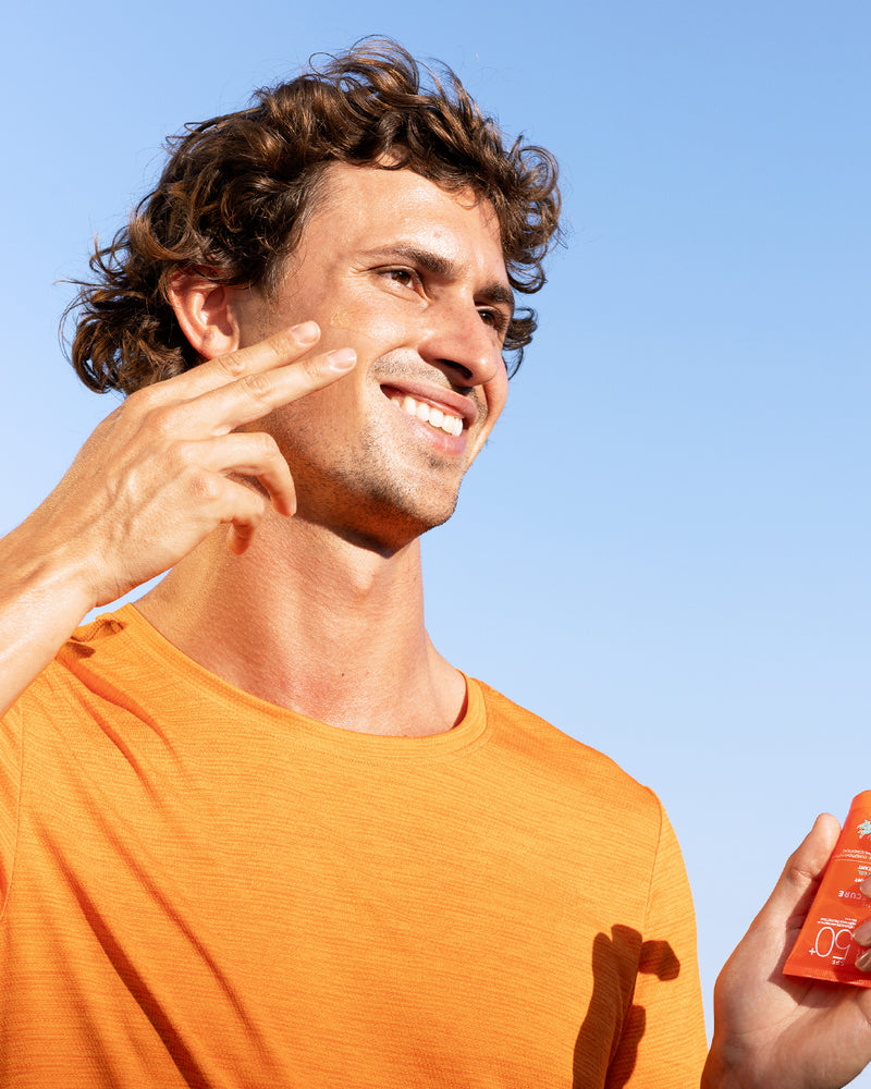 Mann in orangefarbenem Shirt trägt SUN SECURE Extrême SPF50+ Gel auf sein Gesicht auf, vor blauem Himmel.