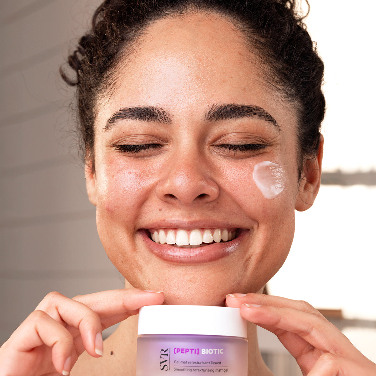Smiling woman applying SVR [PEPTI] Biotic smoothing matt gel from a purple jar to her cheek.