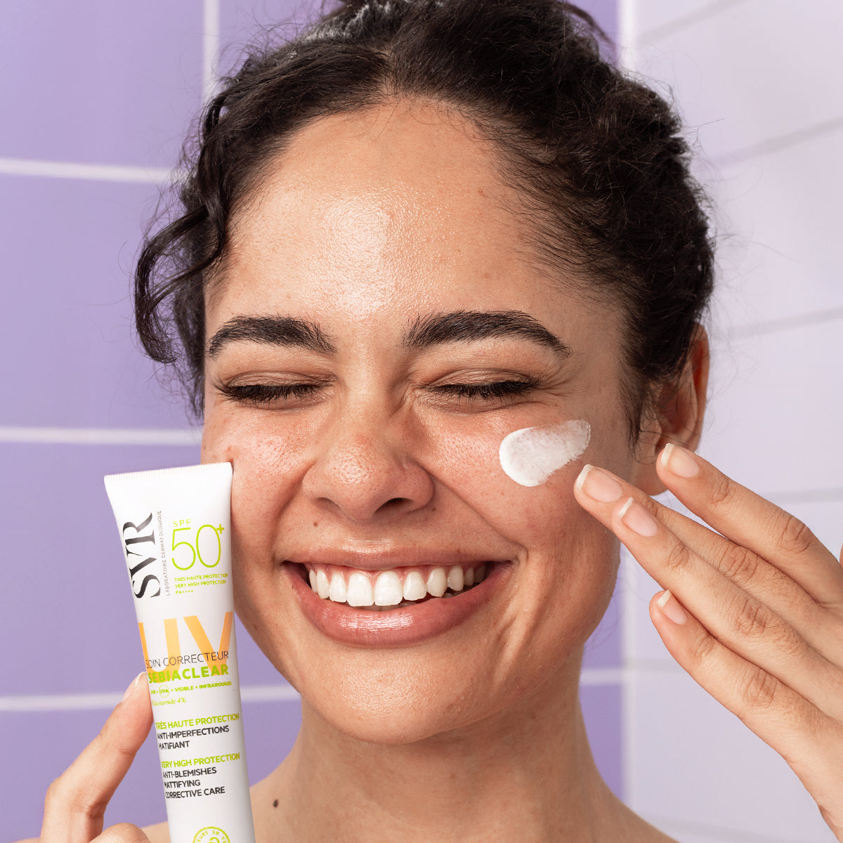 Frau trägt SVR Sebiaclear SPF50+ Creme auf ihre Wange auf, vor einem hellvioletten Fliesen-Hintergrund.