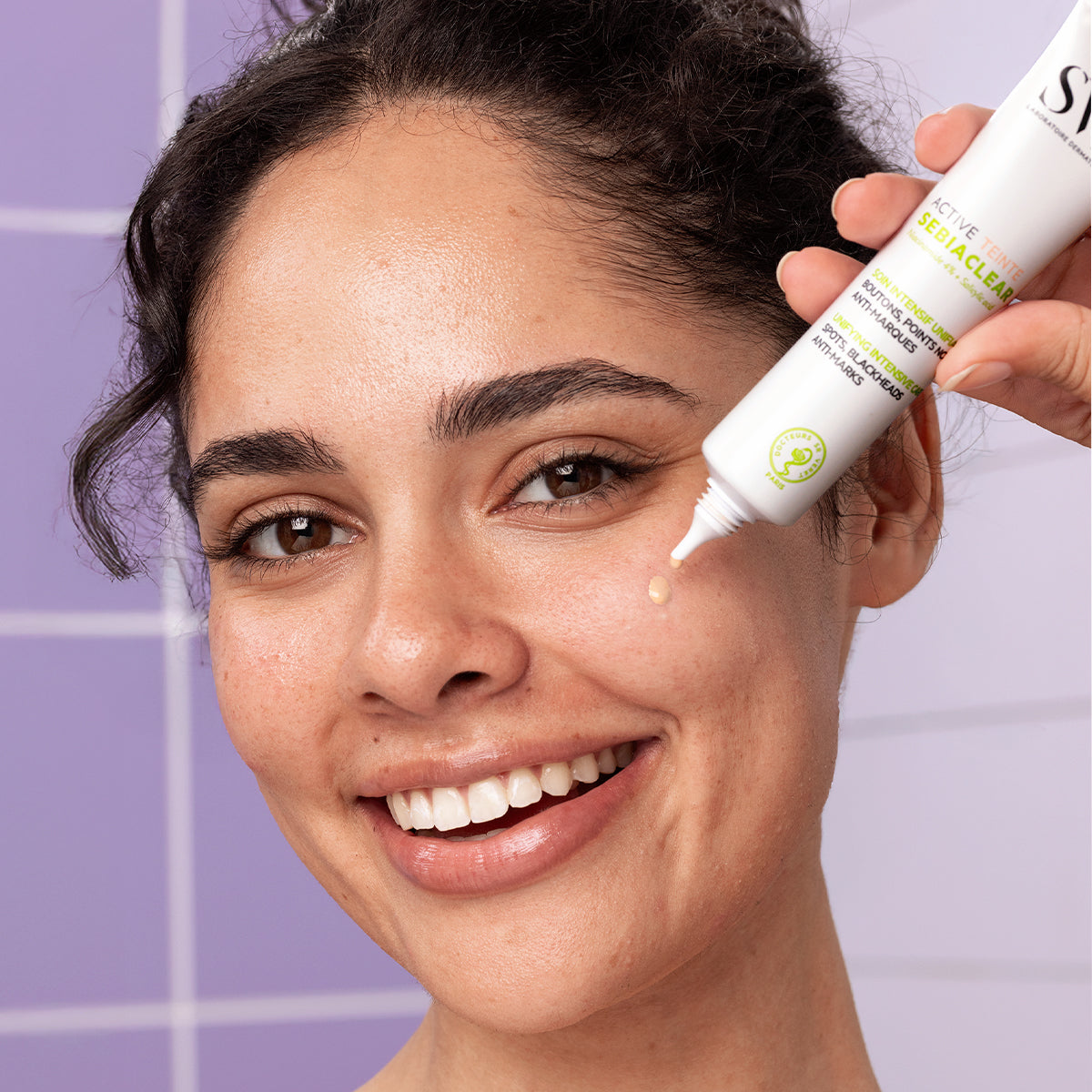 Frau trägt SVR Sebiaclear Active Teinté Creme auf ihre Wange auf, vor einem lila gekachelten Hintergrund.