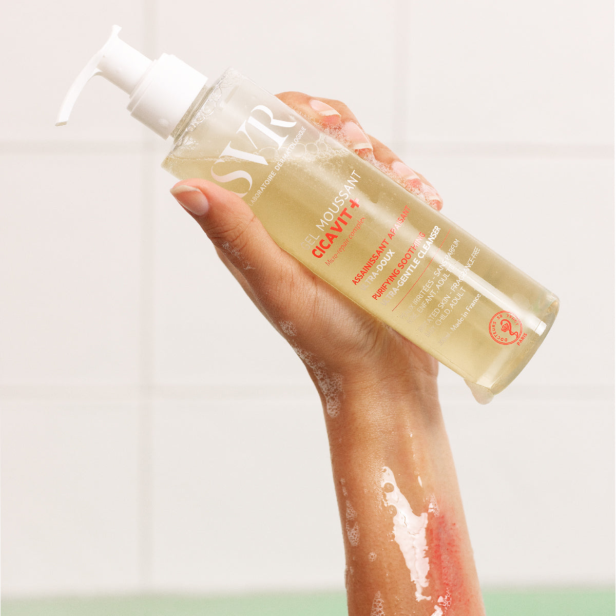 Hand hält eine transparente SVR CICAVIT+ Gel Moussant Pumpflasche vor einem weißen Fliesen-Hintergrund.