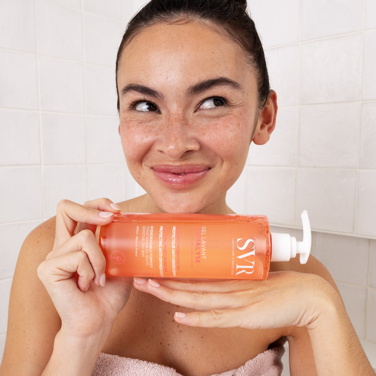 Frau hält eine orangefarbene SVR TOPIALYSE Gel Lavant Pumpflasche vor weißem Fliesen-Hintergrund.
