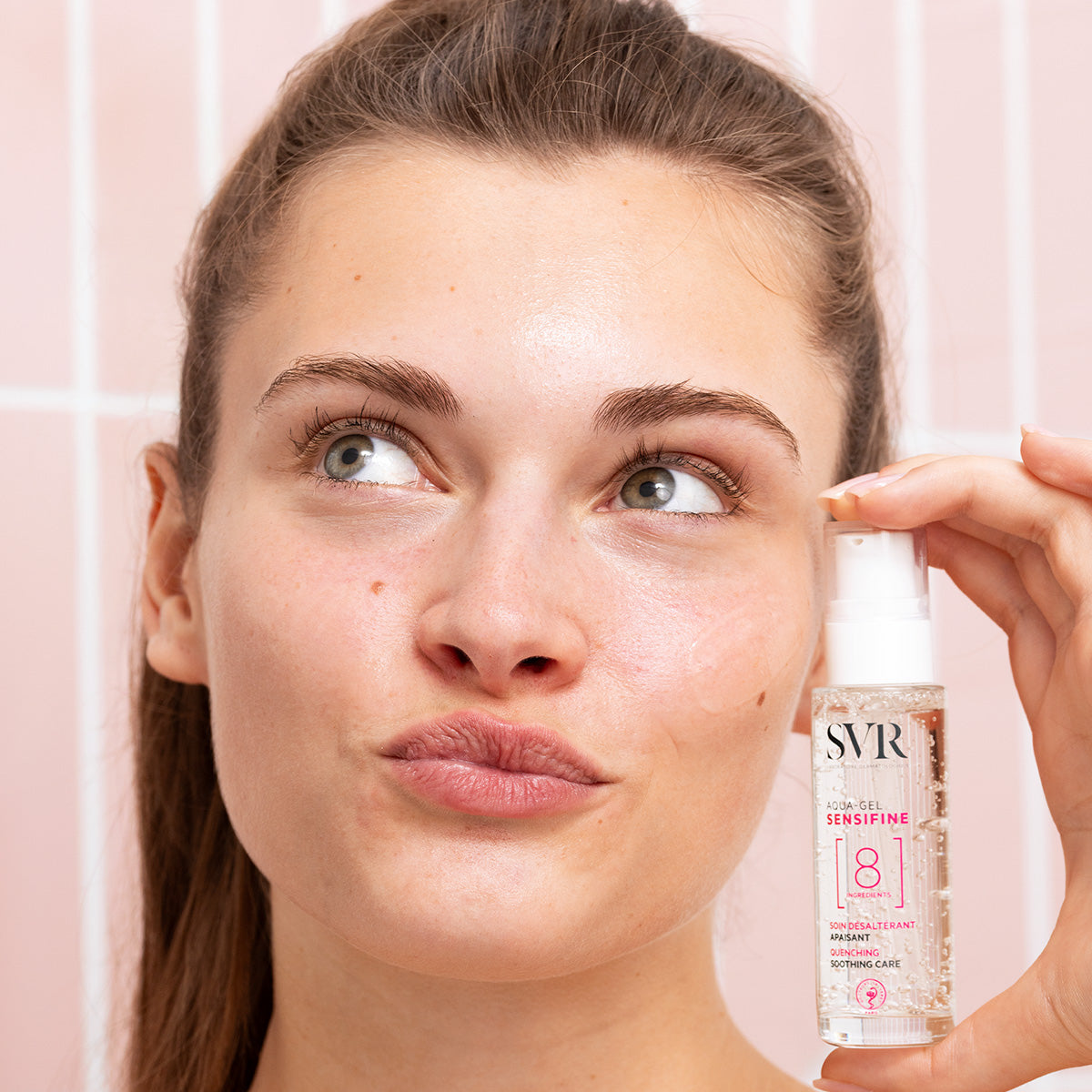 Frau hält SVR Sensifine Aqua-Gel Pumpspender mit transparenter Geltextur vor rosa gekacheltem Hintergrund.