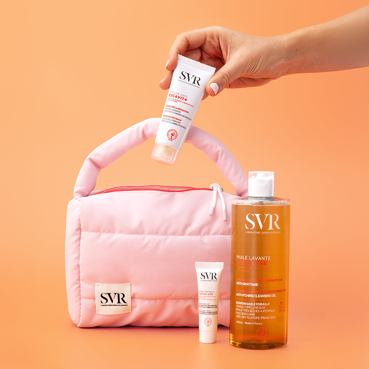 Hand hält SVR Cicavit+ Creme über einer rosa gesteppten Kosmetiktasche, daneben SVR Topialyse Reinigungsöl und kleine Tube.
